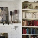 A partial view of a tall white built-in shelf with books groups more or less by color. To the left is a fireplace mantel with blue candles.