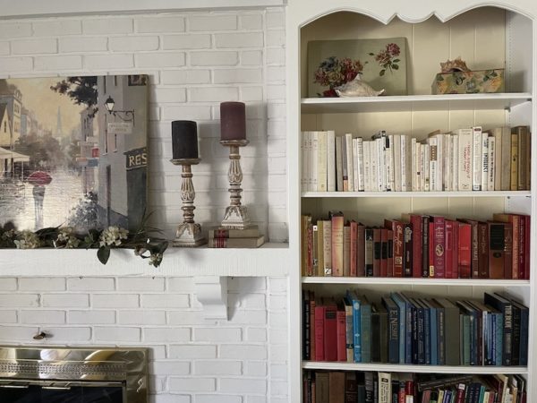 A partial view of a tall white built-in shelf with books groups more or less by color. To the left is a fireplace mantel with blue candles.